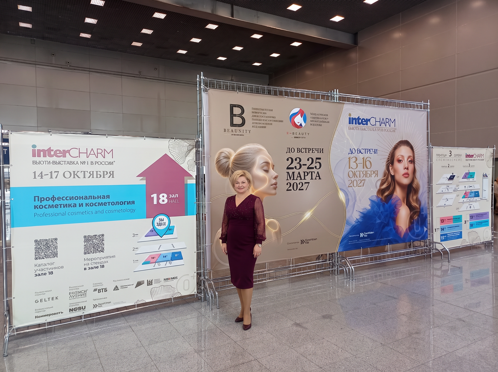 Женщина в бордовом платье стоит на фоне баннеров выставки InterCHARM с датами мероприятий и информацией о профессиональной косметике и косметологии